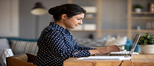 Young adult indian woman student using laptop computer study in internet, focused girl typing looking at notebook working online e learning with education software technology sit at home office table